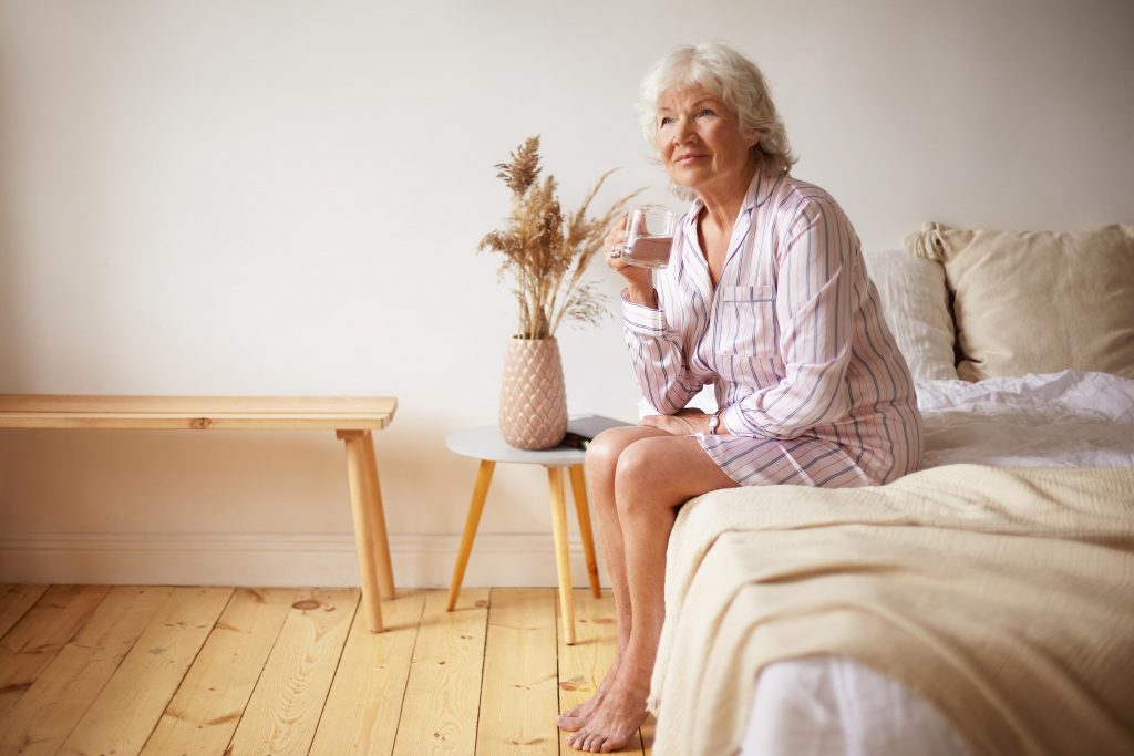 indoor shot barefooted attractive gray haired female pensioner sitting bed with feet wooden floor holding glass drinking fresh water morning people lifestyle bedtime aging concept