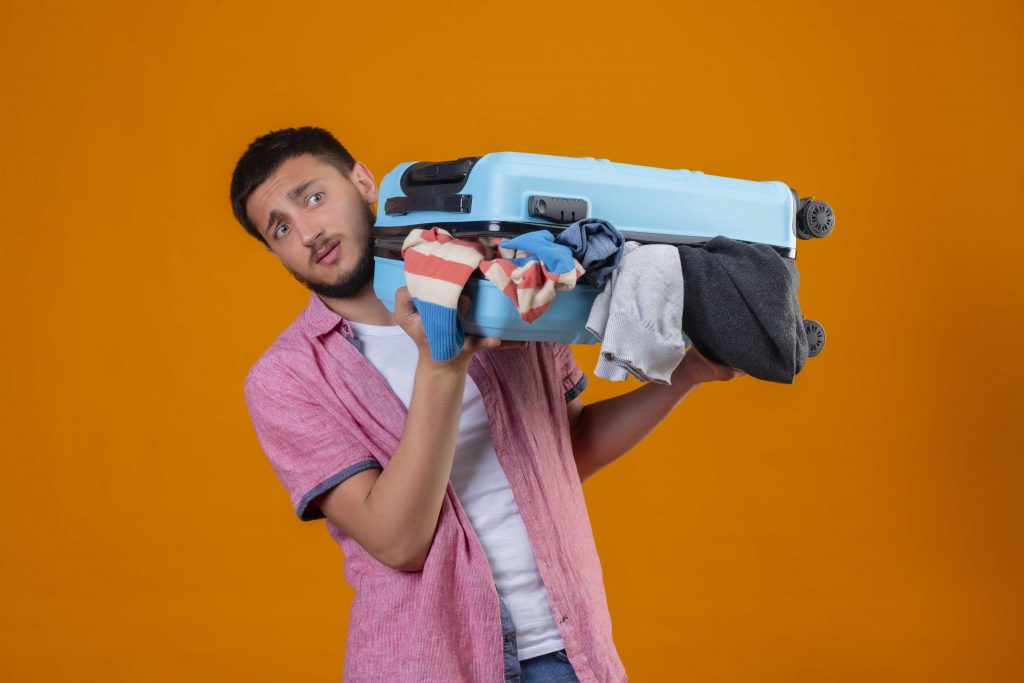 upset young handsome traveler guy holding suitcase full clothes looking aside with sad expression standing orange background